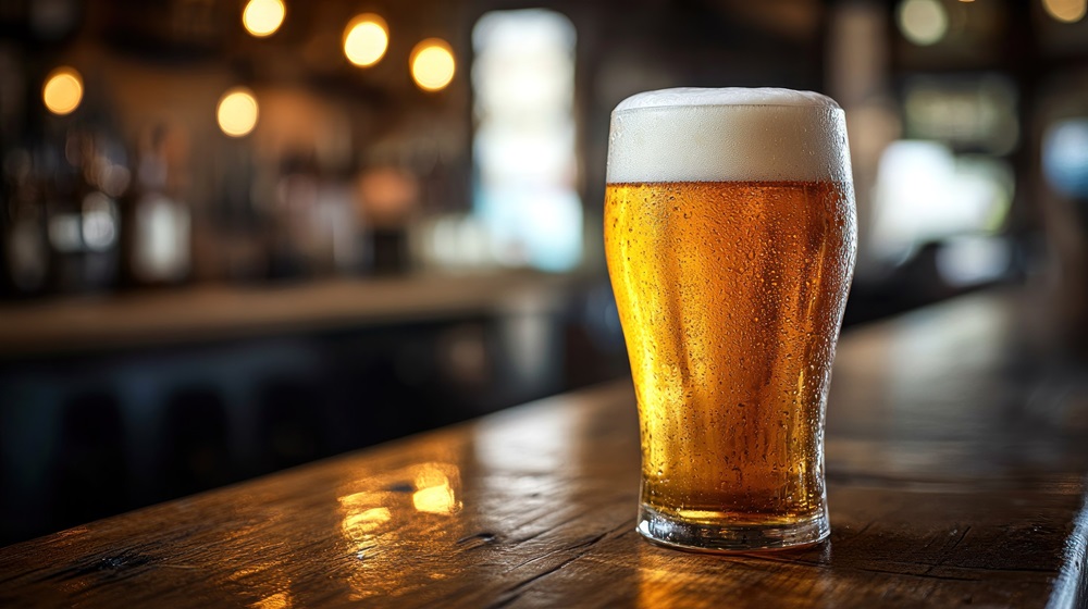 Ein Glas goldenes Bier mit Schaum steht auf einer dunklen Holztheke; im Hintergrund sorgen warme, unscharfe Lichter für eine gemütliche Atmosphäre.