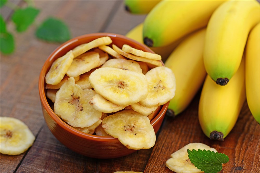 Bananenscheiben in einer Schale vor frischen Bananen auf Holztisch.