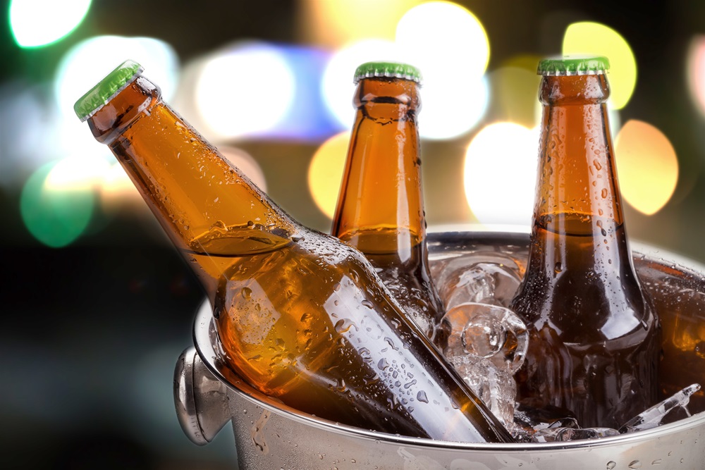 Nahaufnahme mehrerer brauner Bierflaschen mit grünen Kronkorken, die von links nach rechts schräg angeordnet sind; im Hintergrund leuchten bunte, stark verschwommene Lichter.