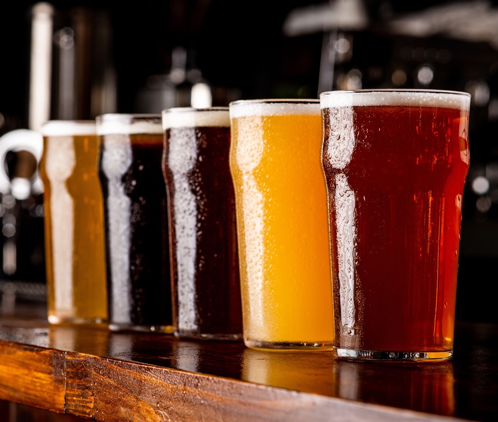 Nahaufnahme mehrerer Gläser mit unterschiedlich gefärbtem Bier und Schaum auf einer dunklen Holzoberfläche; im Hintergrund sind weitere unscharfe Biergläser zu sehen.
