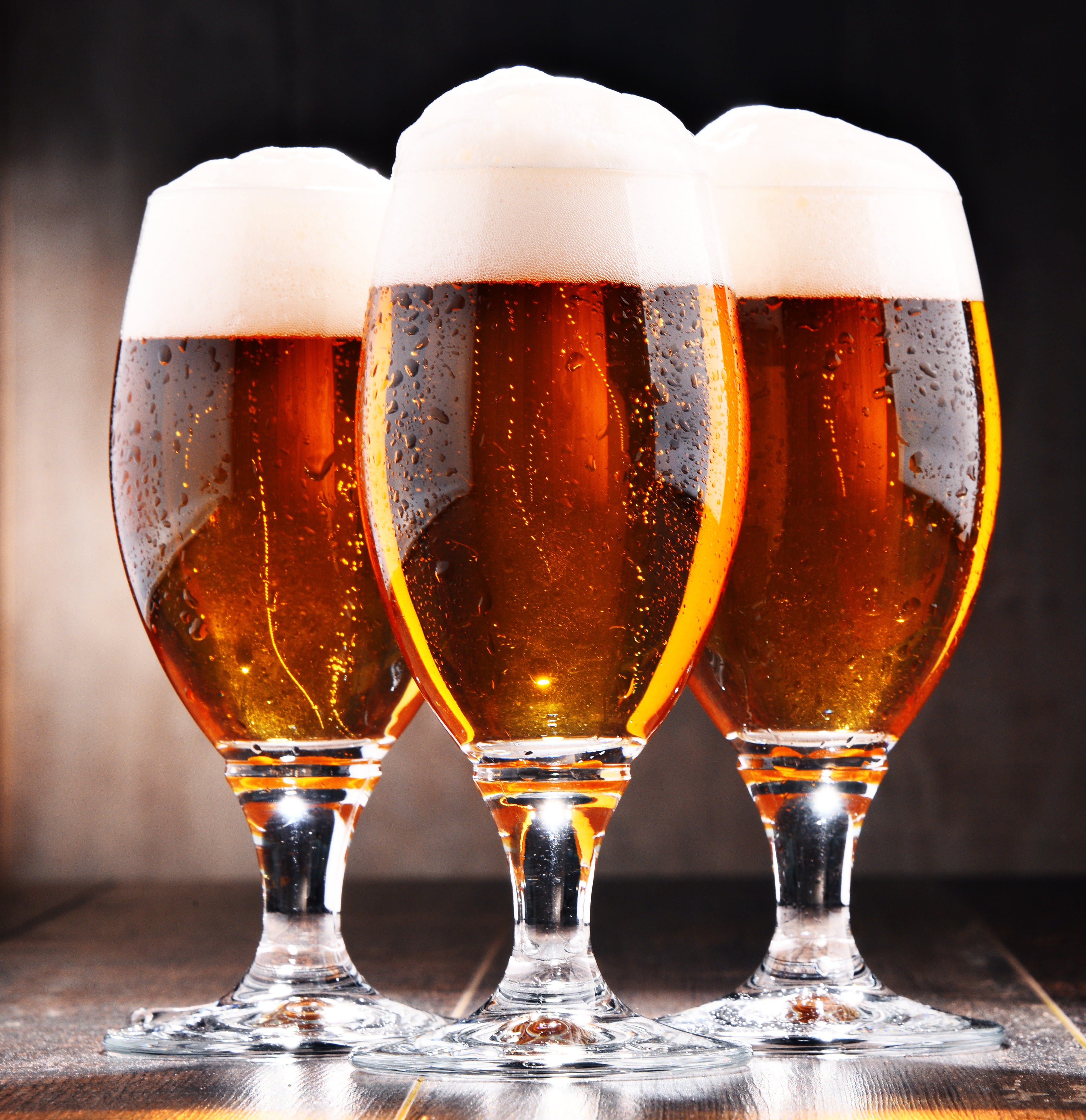 Drei Biergläser mit Bier und Schaum, die auf einer hölzernen Oberfläche nebeneinander stehen, mit einem unscharfen Hintergrund, der eine gemütliche Atmosphäre vermittelt.