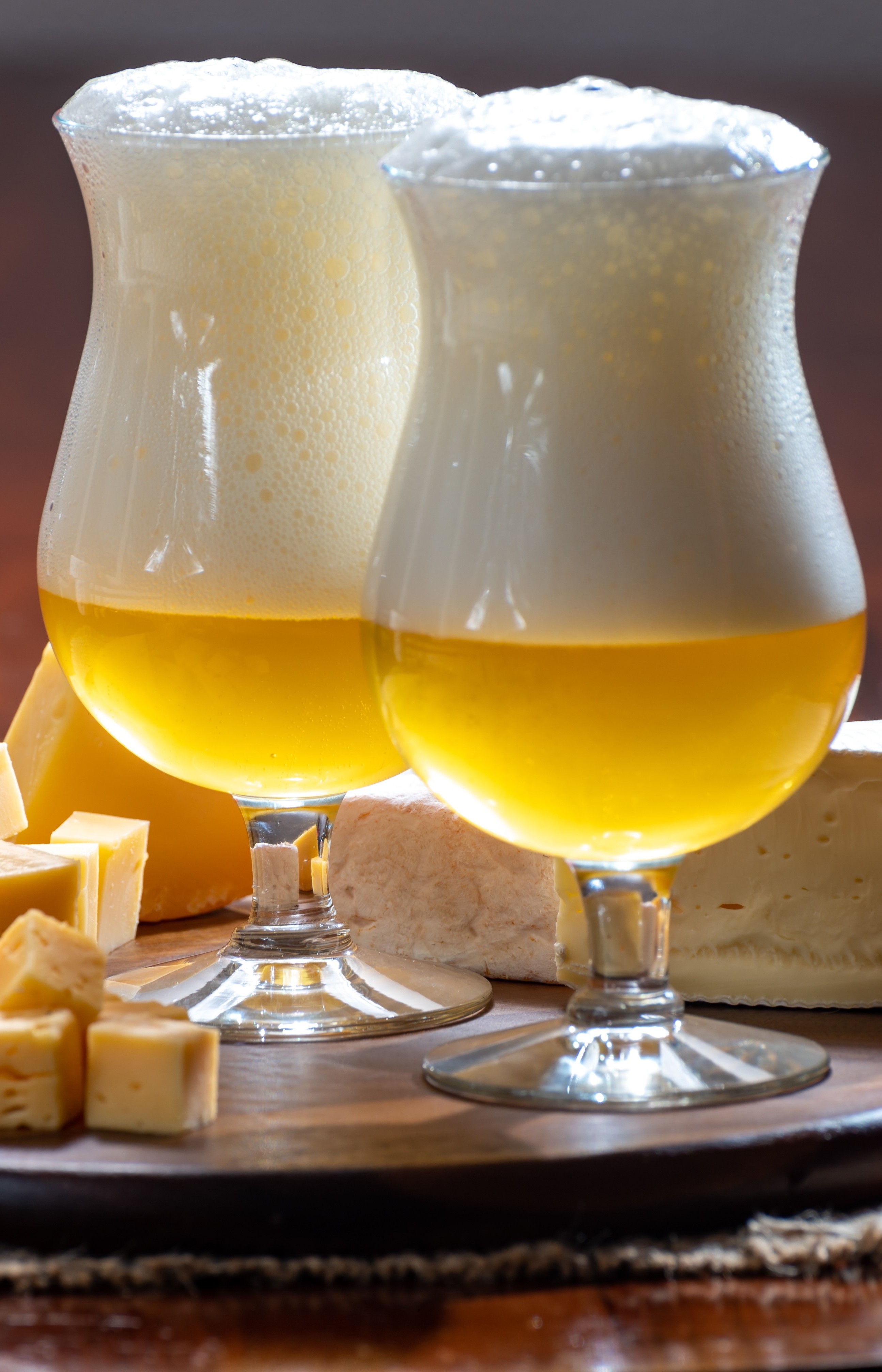 Zwei Gläser Bier mit üppiger Schaumkrone stehen auf einem runden Holzbrett, umgeben von verschiedenen Käsesorten wie Würfeln, Scheiben und einem Stück Weichkäse.