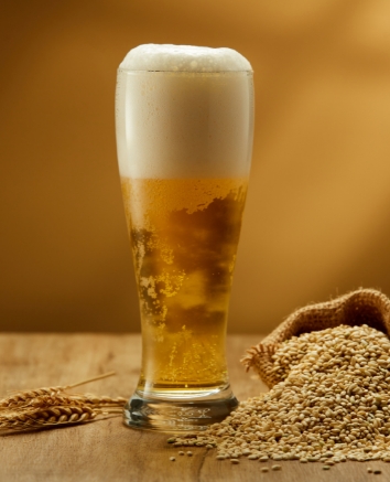 Ein Glas frisch gezapftes, goldgelbes Weißbier mit dichter Schaumkrone auf einem Holztisch, daneben ein Sack mit Gerstenmalz.